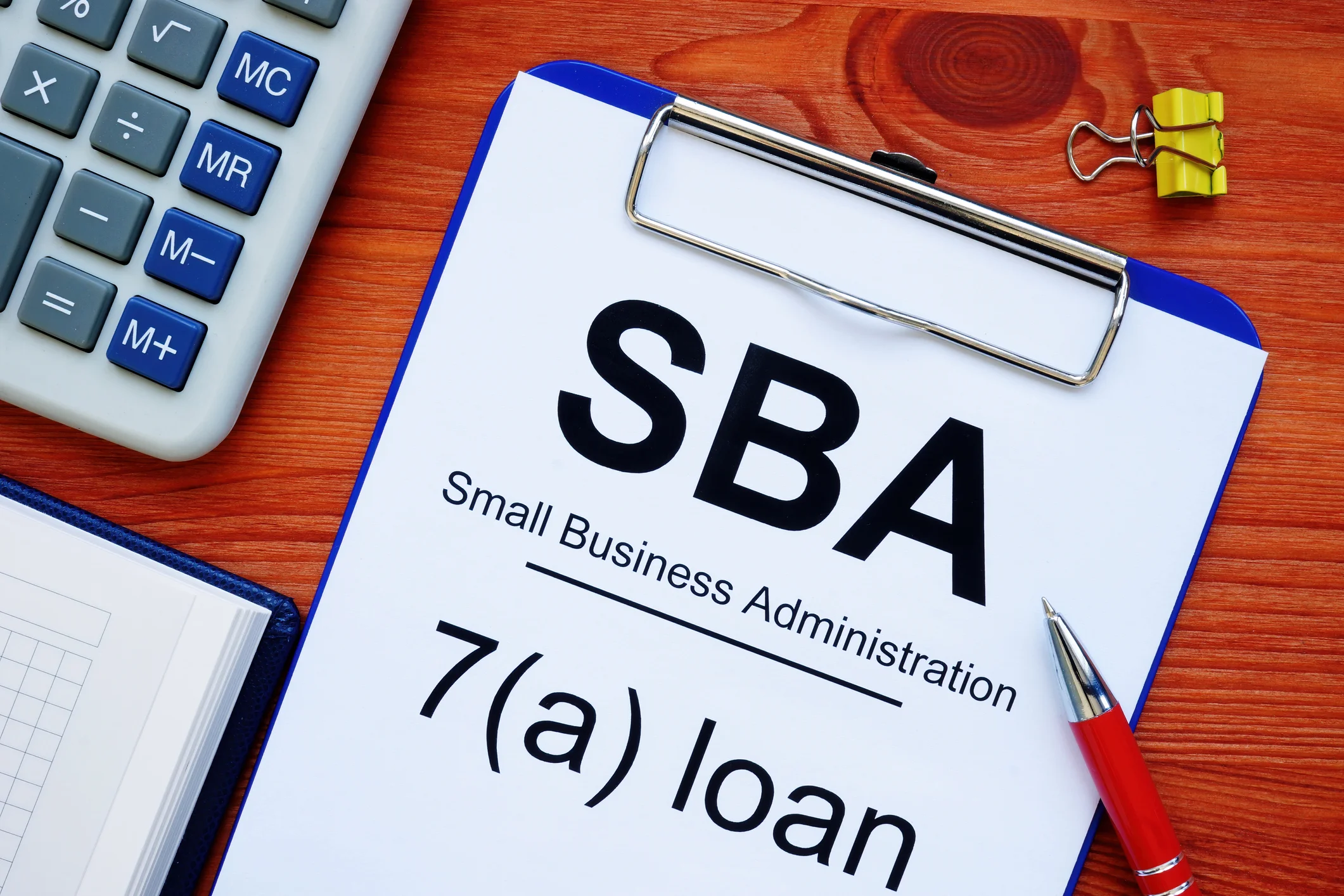 Clipboard with SBA Small Business Administration 7(a) loan paperwork on a wooden desk next to a calculator, notepad, and pen — representing SBA loan options for government contractors seeking working capital and contract financing