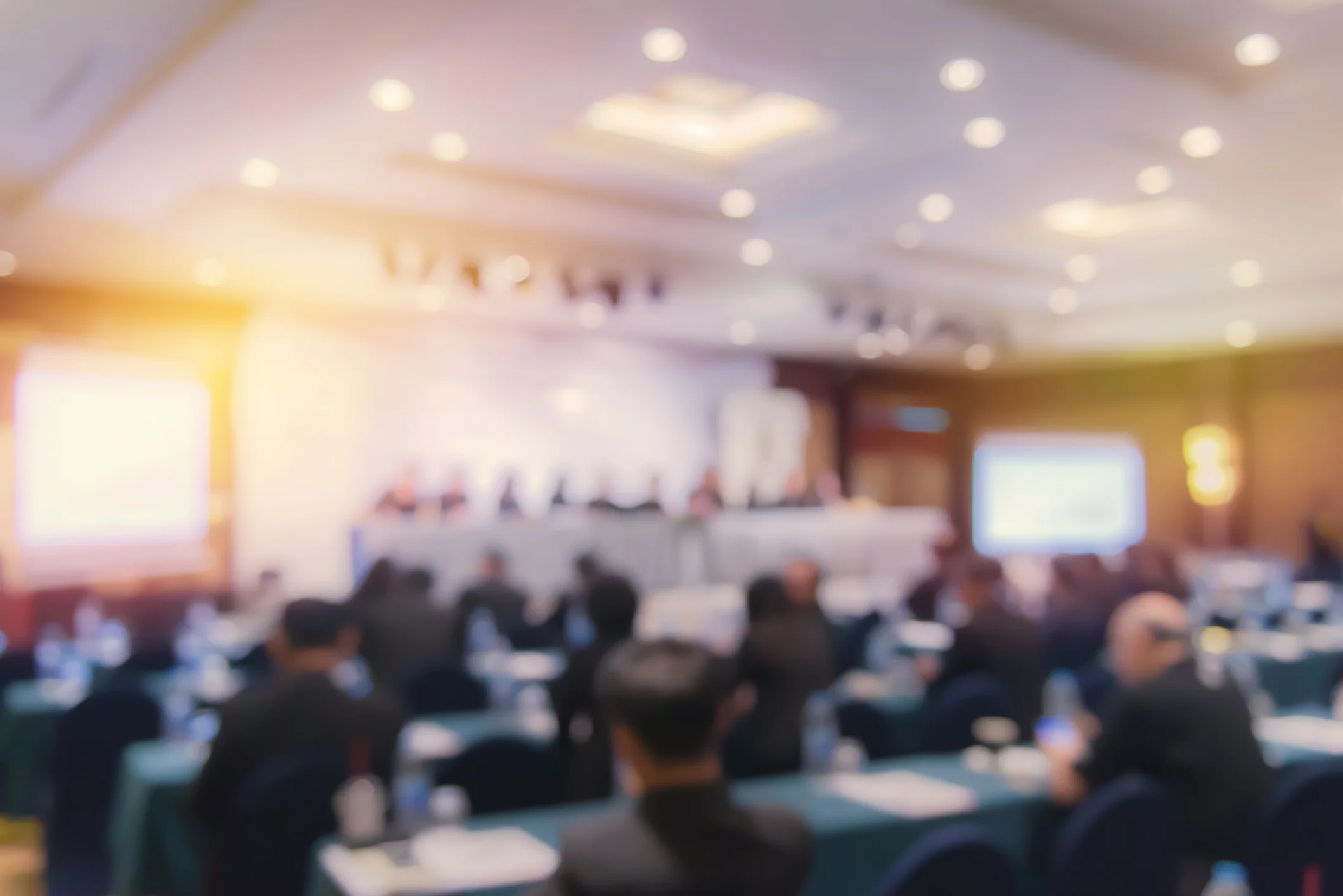Blurred photo of a professional government contracting conference showing attendees seated at tables facing a speaker panel on stage with warm lighting